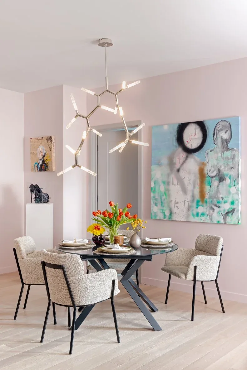Pale pink dining room with a round dark table, X-shaped base, light textured chairs and a slim branching pendant