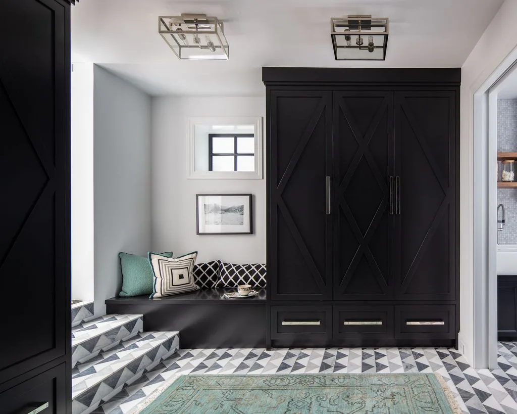 Black mudroom cupboards with diagonal door details, lower drawers, dark bench, patterned cushions, black-and-white geometric floor, and pale walls.