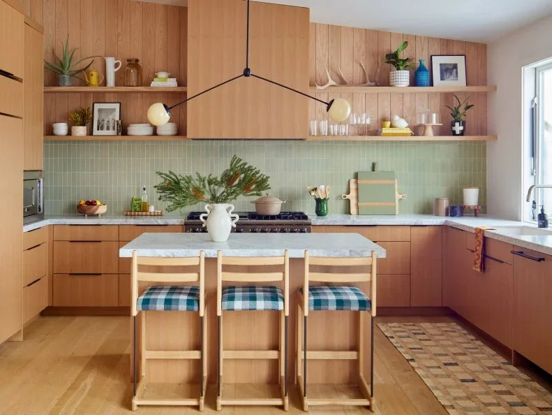 White oak cabinets and open shelves surround pale green vertical tiles, with checked stool cushions at a small island.