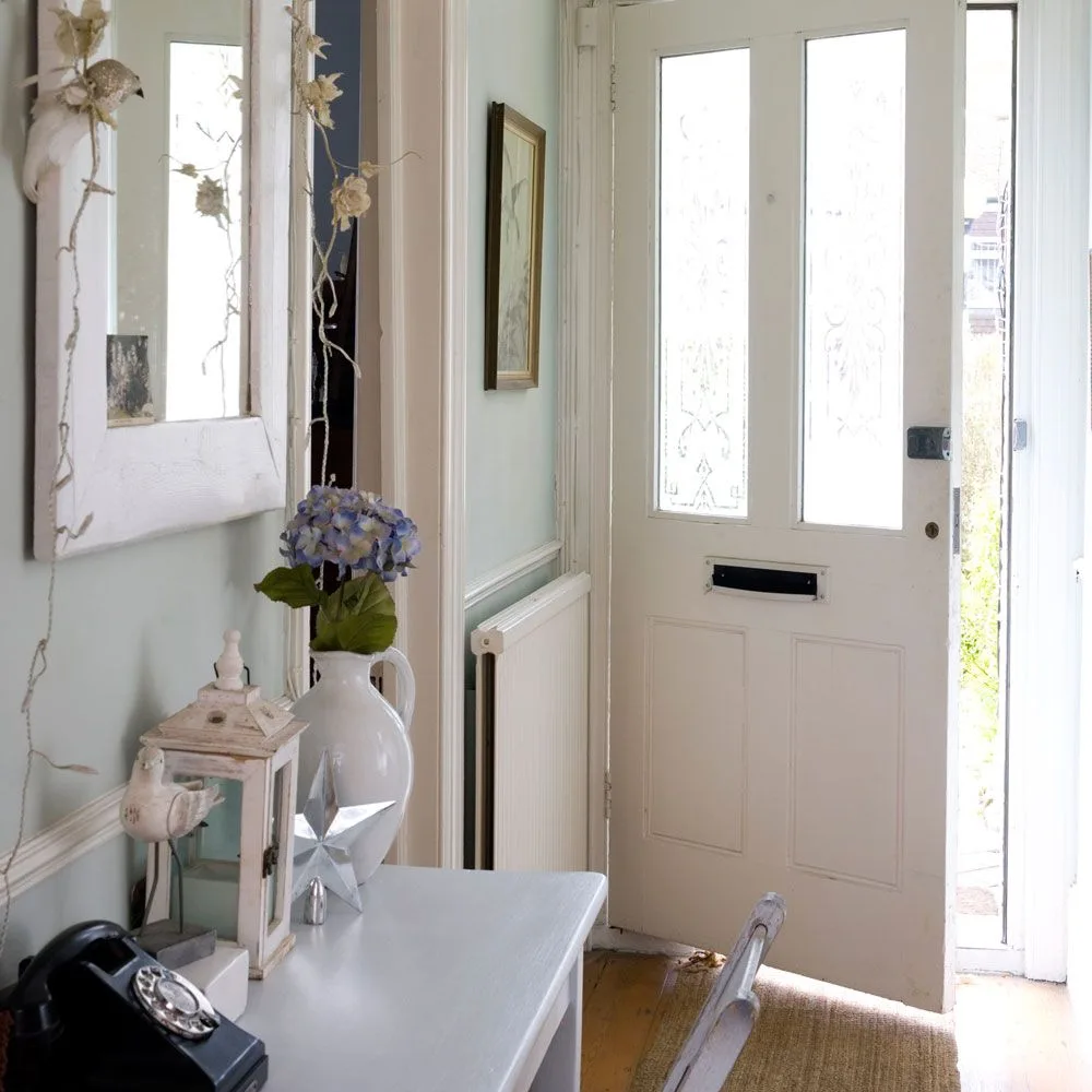Pale entry table with white pitcher of blue flowers, small lantern, black telephone and mirror beside a glazed white door