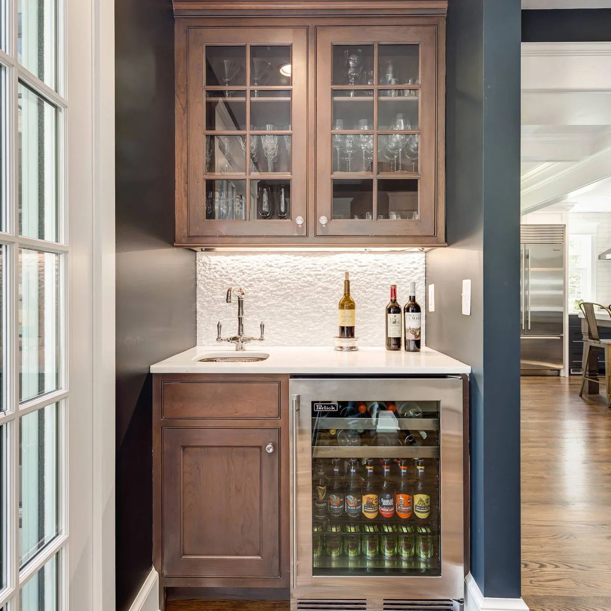 Small wood mini bar with glass-front upper cupboard, pale textured backsplash, sink and beverage refrigerator