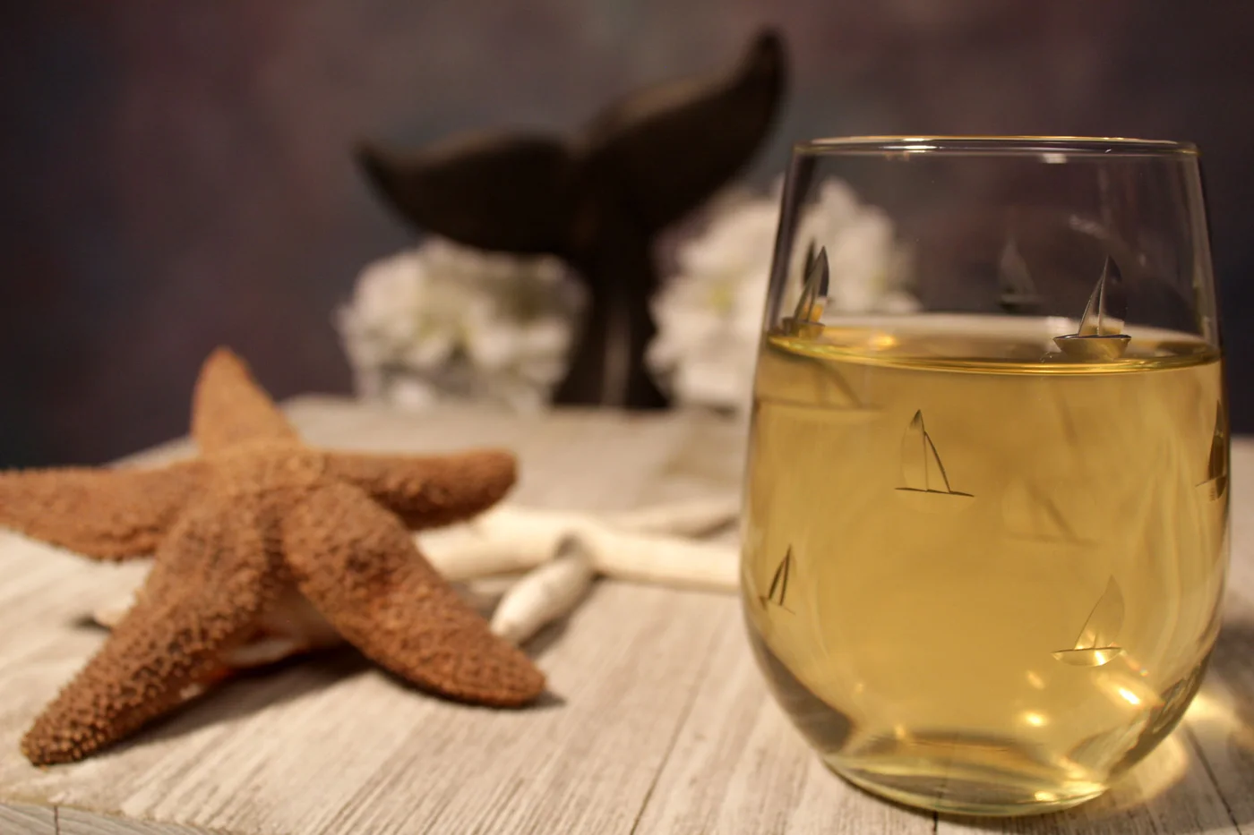 A clear stemless glass with etched sailing motifs stands beside pale dishes and a starfish decoration on a wood surface