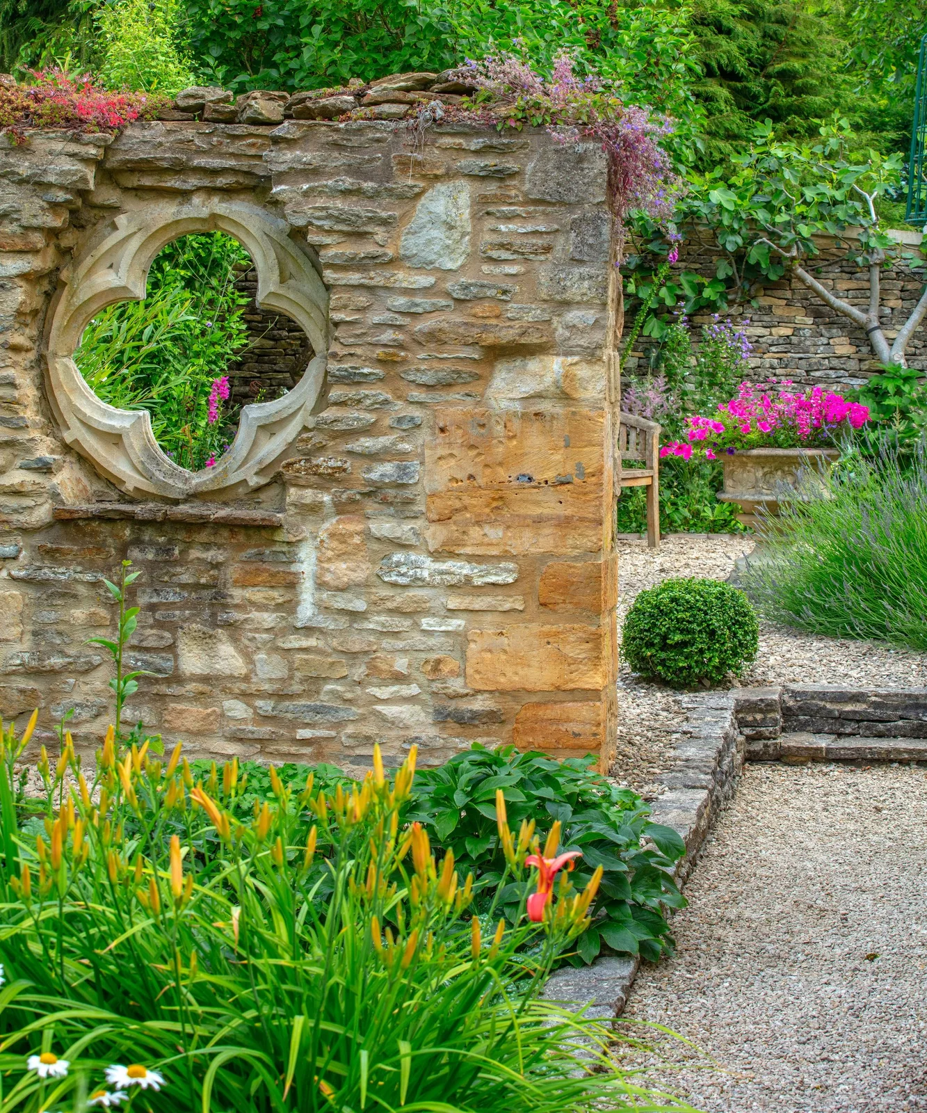 A stone garden wall with a shaped opening partially screens a bench and planted gravel area