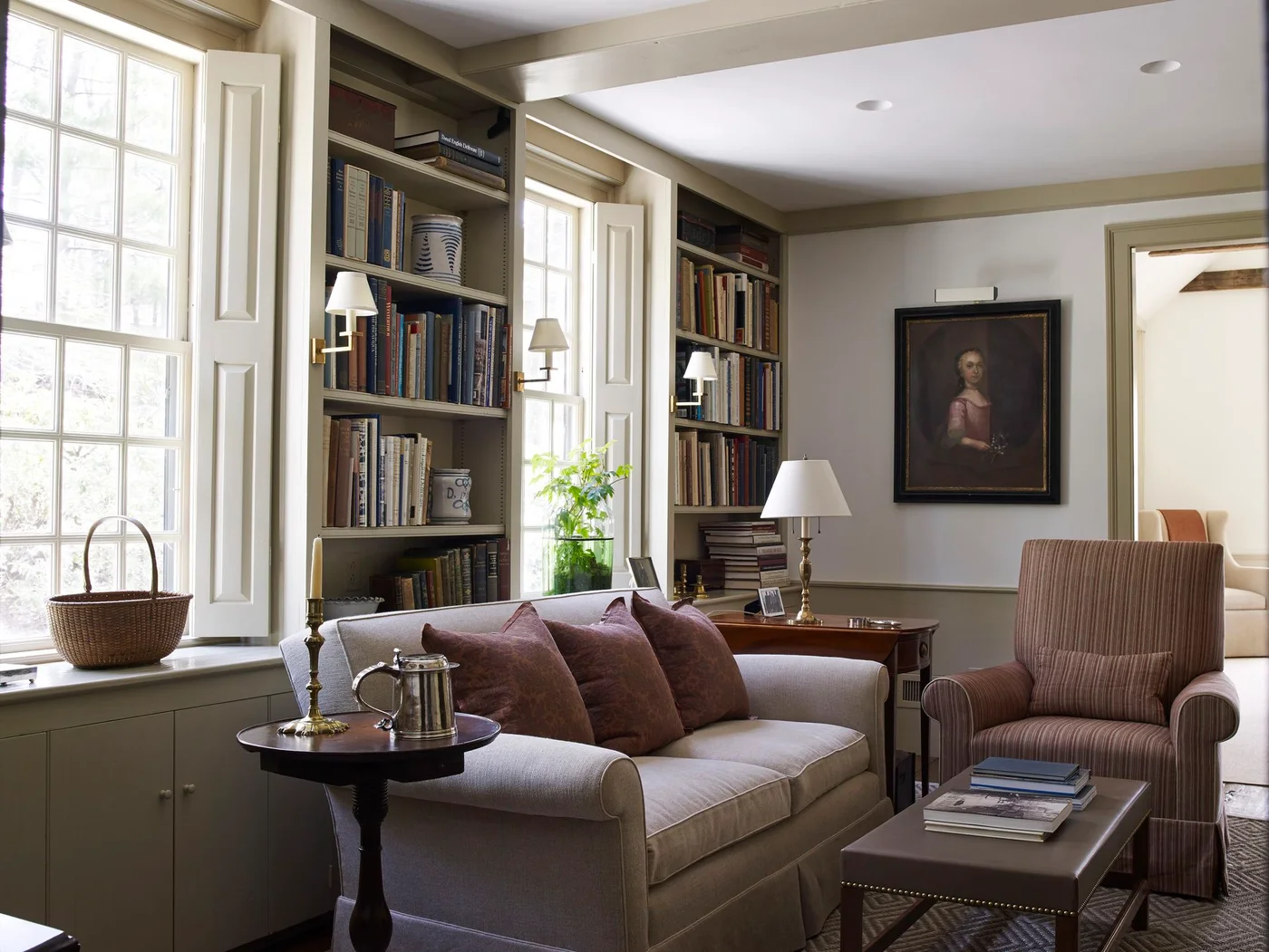 Bookcases sit between tall shuttered windows behind a pale sofa, striped armchair and several small reading tables