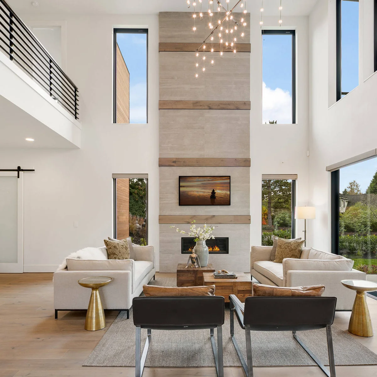 A double-height living room with a tall pale fireplace wall crossed by wood bands, facing white sofas and a hanging light cluster.