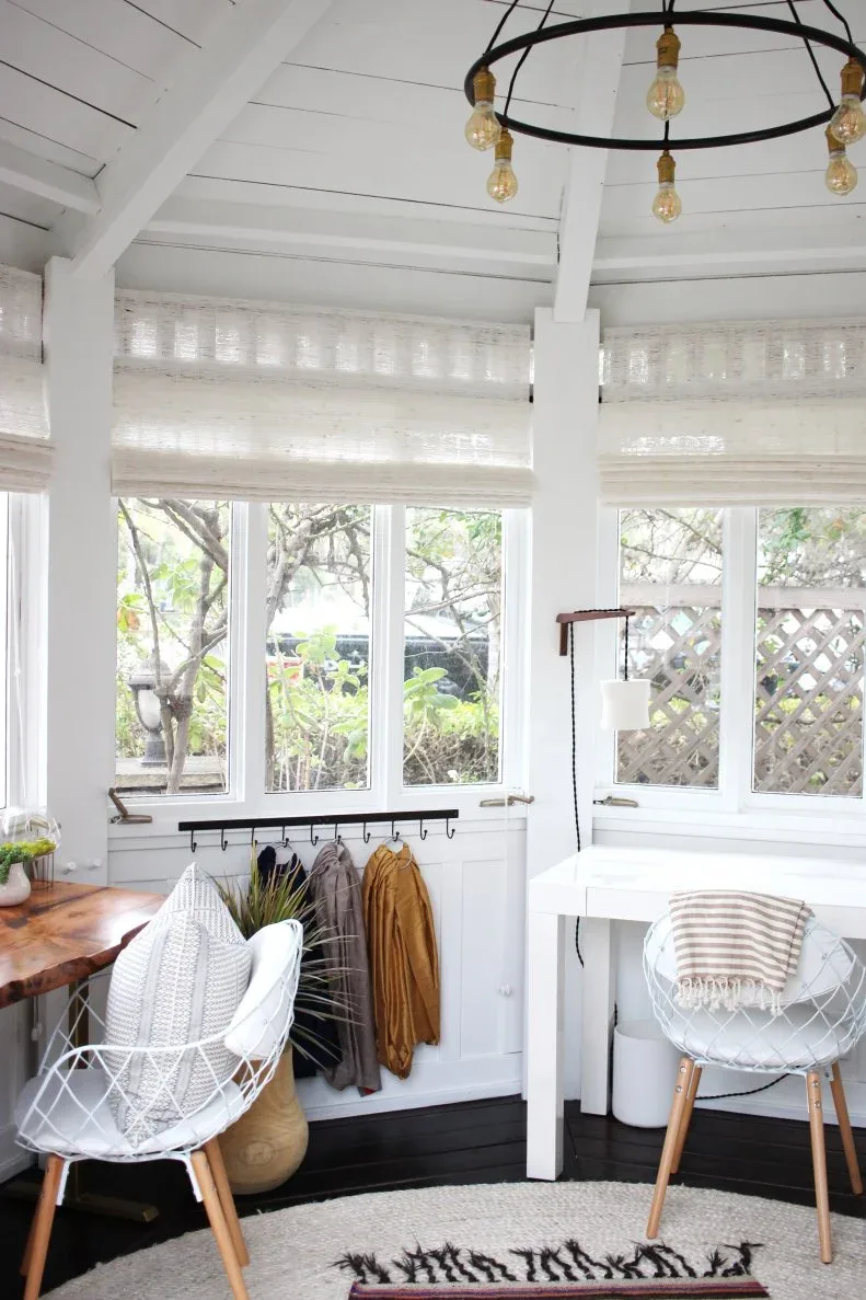 A bright glazed workspace has separate white and wood desks, white open-frame chairs, individual lamps, pale window shades and a rail of hooks between the work areas.