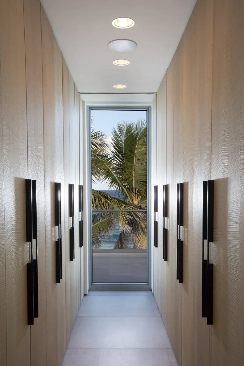Pale full-height closet doors with long black handles line a narrow passage ending at a window.