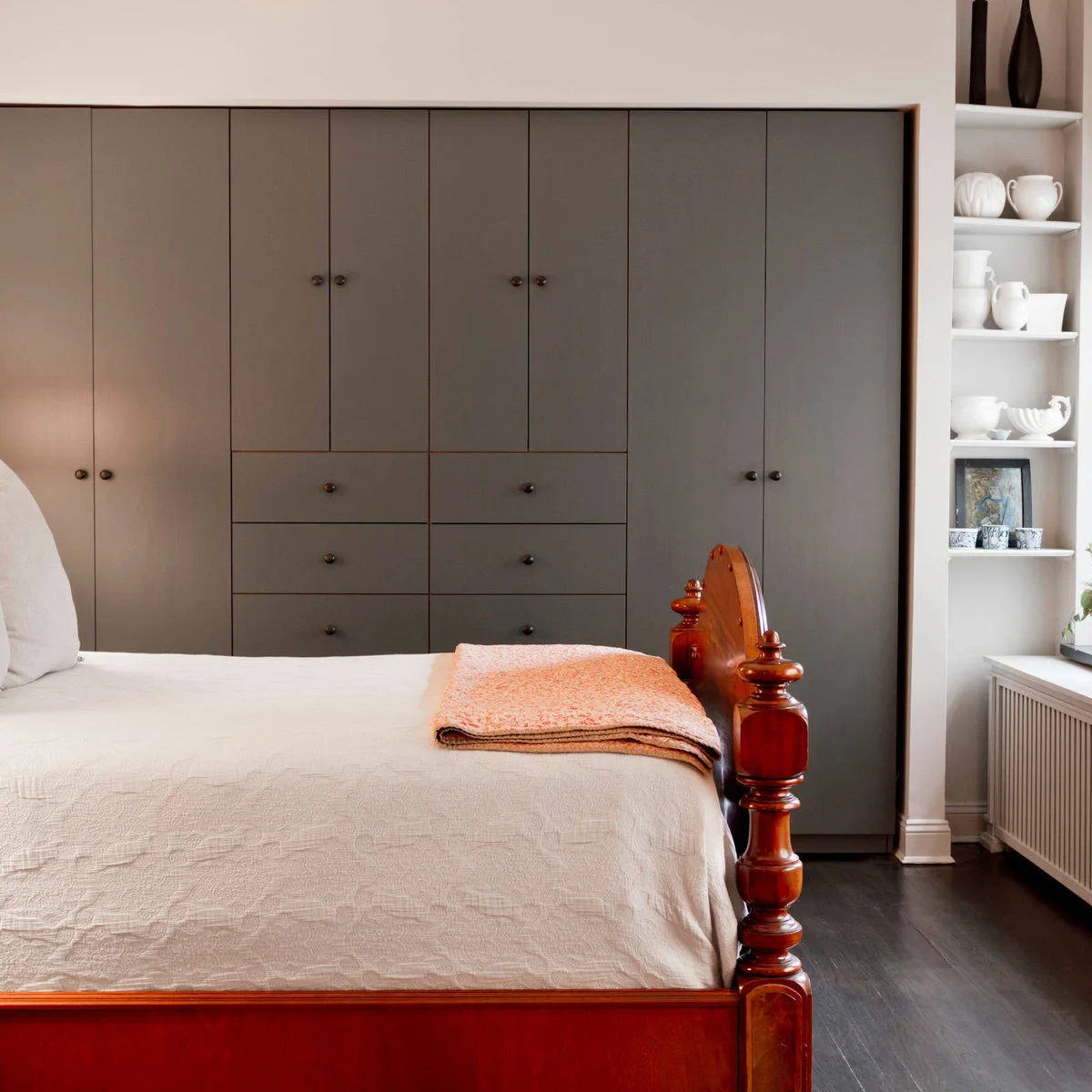 Bedroom with a gray wall of fitted cupboards and drawers beside a traditional wood bed