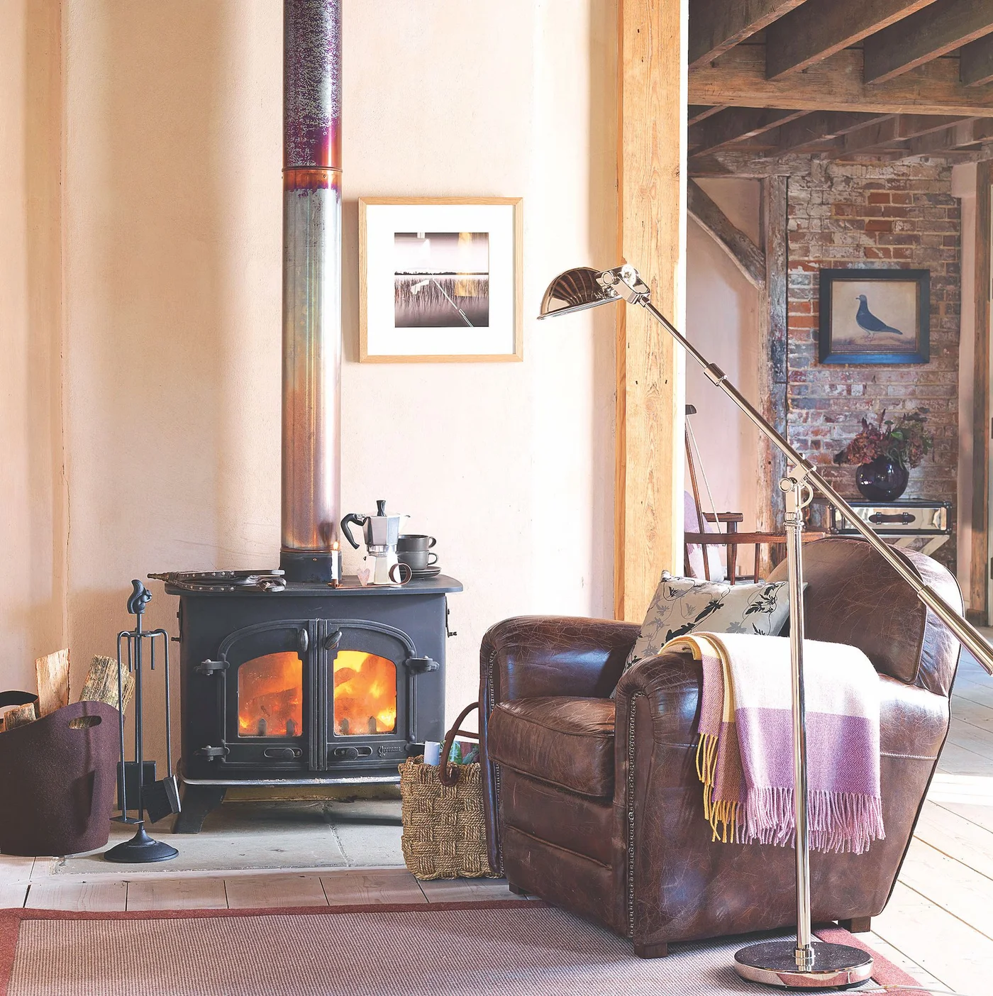 A brown chair sits beside a dark stove, exposed wooden framing, brick walls and a slender reading lamp