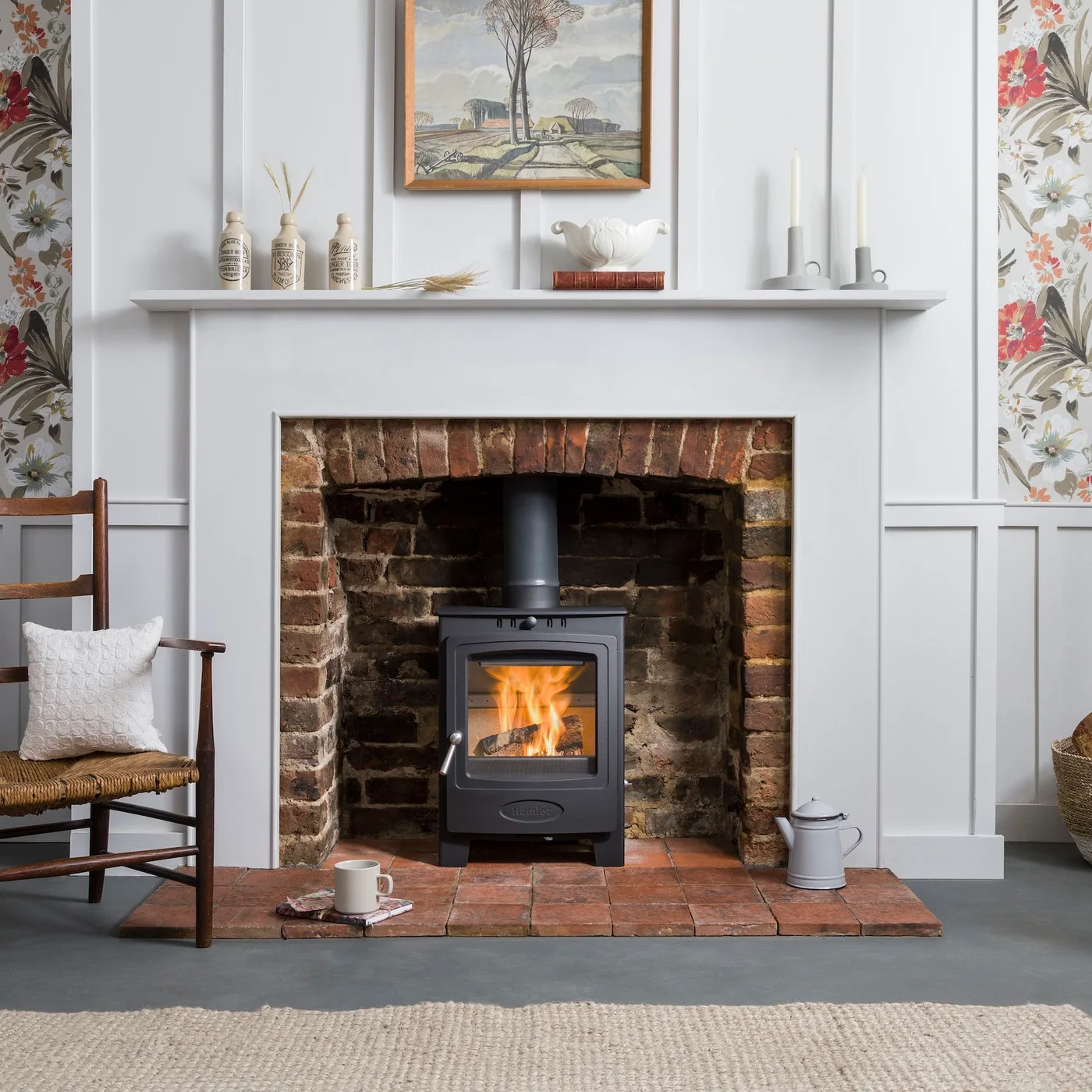 A black stove sits inside an exposed brick opening on a reddish tiled hearth beneath a pale paneled mantel and floral wallpaper.