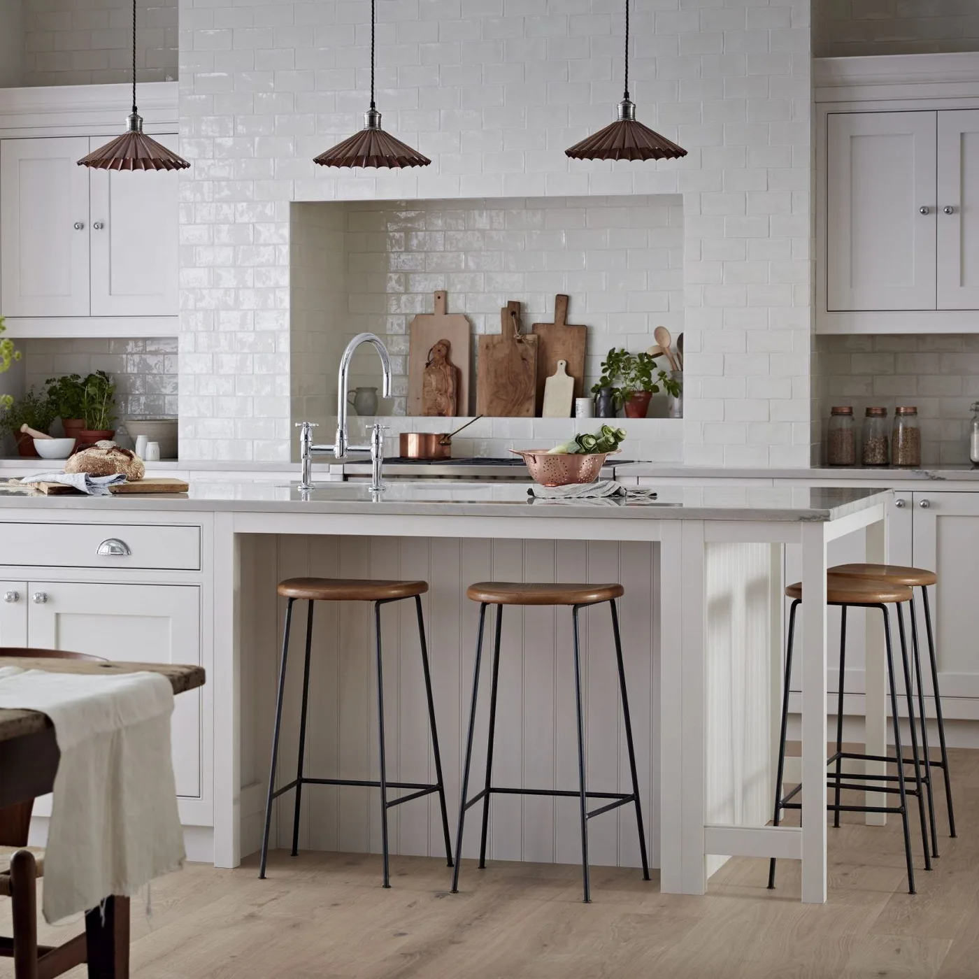 White kitchen with tiled cooker alcove, paneled seating island, three dark-framed stools and small shaped pendant shades.