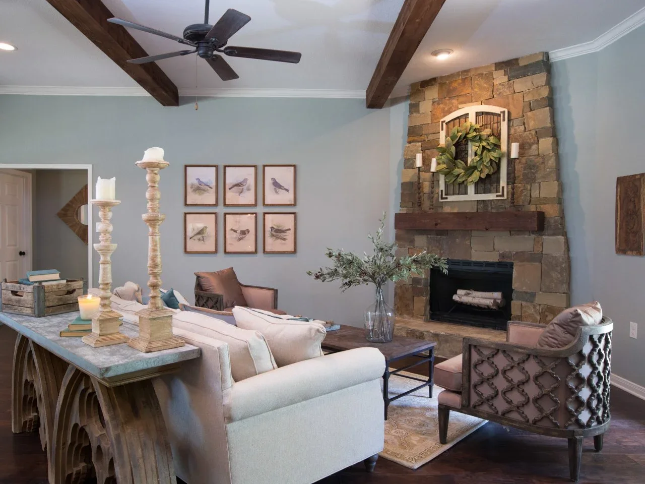A mixed-color stone fireplace and dark timber mantel stand against pale blue walls beneath exposed beams.