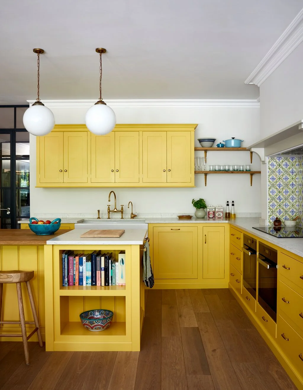 Yellow cabinets and a small peninsula surround a kitchen with floral-patterned tiles at the hob, open glass shelves and a cookbook niche.