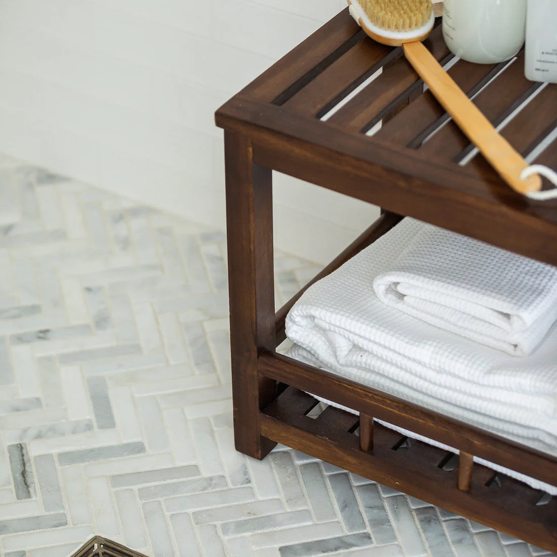 Small pale rectangular mosaic tiles form a herringbone floor beneath a timber bath stool and folded towels.