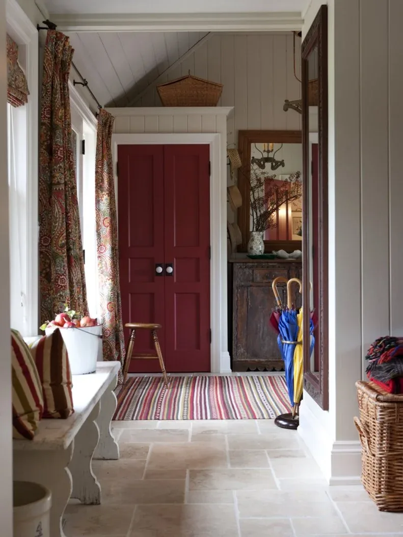 A white freestanding bench with striped cushions sits below windows in a country entry with a red cupboard and patterned curtains.