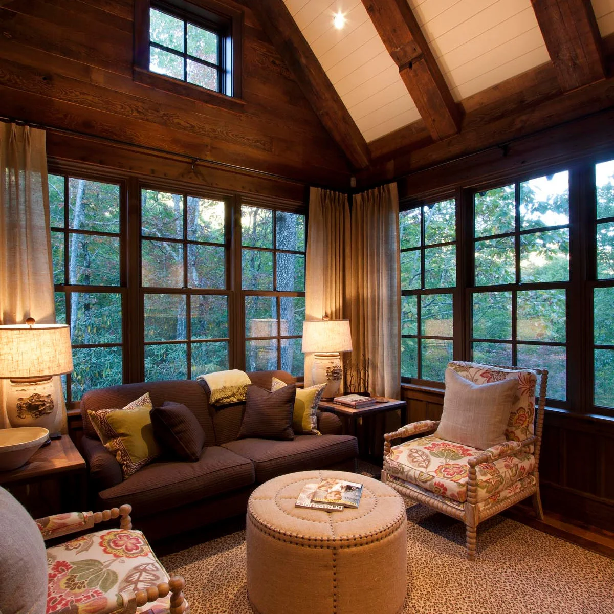 Dark brown fabric sofa with floral chairs, a round tufted ottoman and large lamps in a wood-lined room.