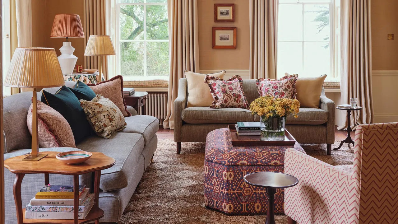 Soft beige seating faces a patterned ottoman beside curtains and a bright window