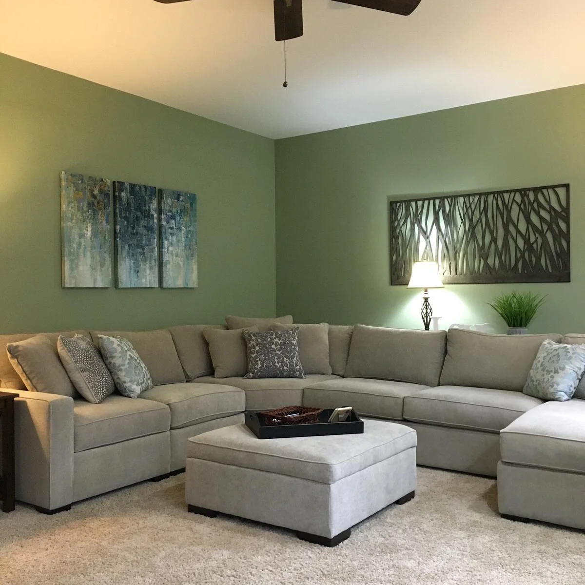 Warm gray sectional and matching ottoman beneath muted sage green walls with gray patterned cushions.