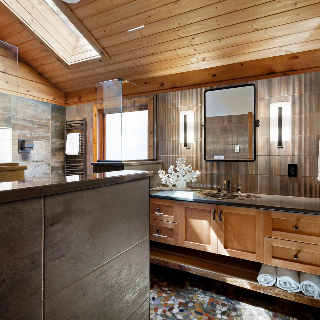 A bathroom with timber ceiling and skylight above a wood vanity, textured tiles and dark framed mirror