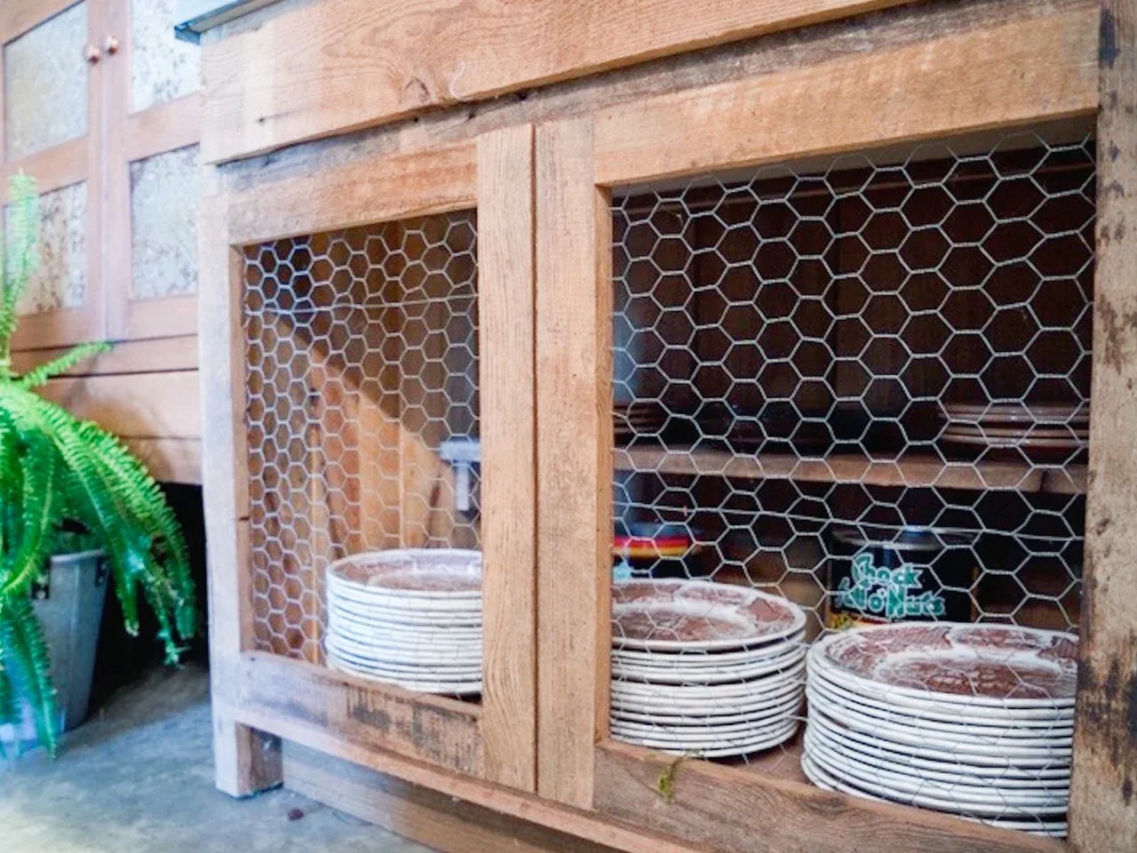 A rustic timber cabinet has wire-mesh doors with stacked patterned plates visible inside.