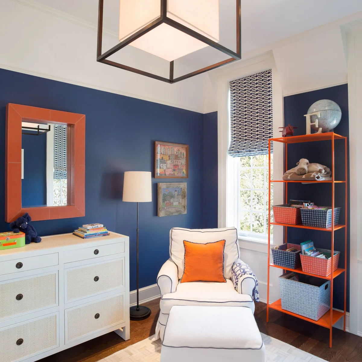 Navy nursery corner with an orange shelf frame, white dresser, pale chair and orange cushion