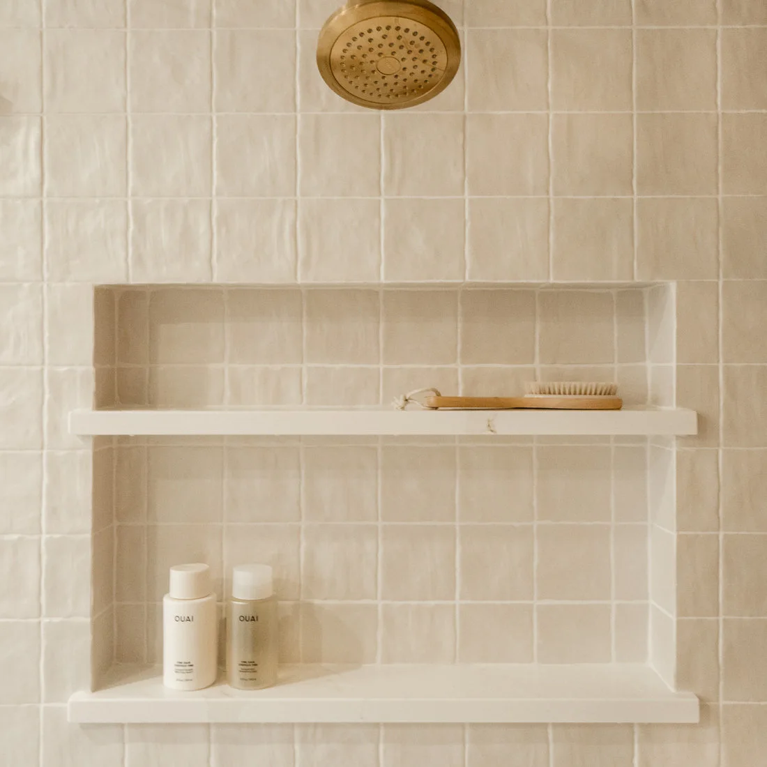 Pale rectangular tiles line two stacked shower niches with light shelf edges beneath a brass-colored showerhead