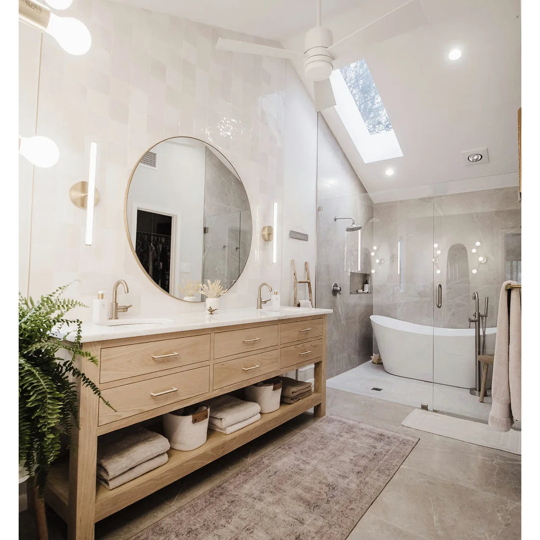 A wood-fronted double vanity and round mirror face pale tiled bathing walls beneath a sloping ceiling and skylight