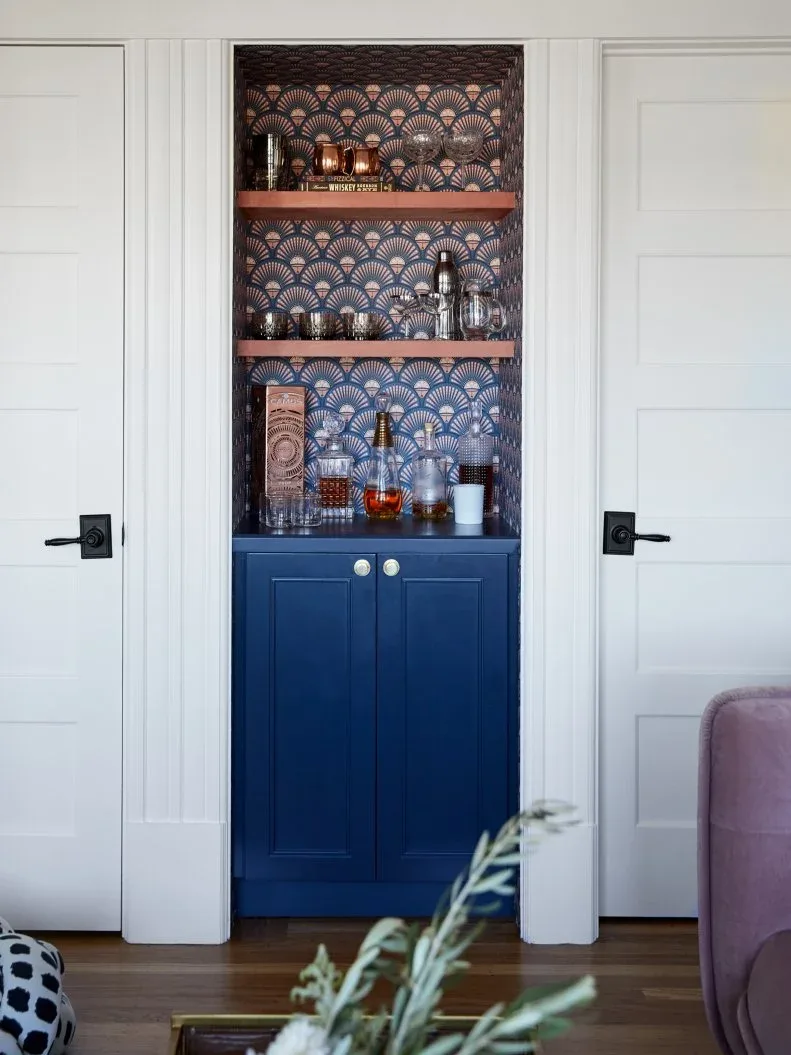 A narrow navy drinks cabinet stands beneath two timber shelves of glassware against patterned wallpaper between white doors.