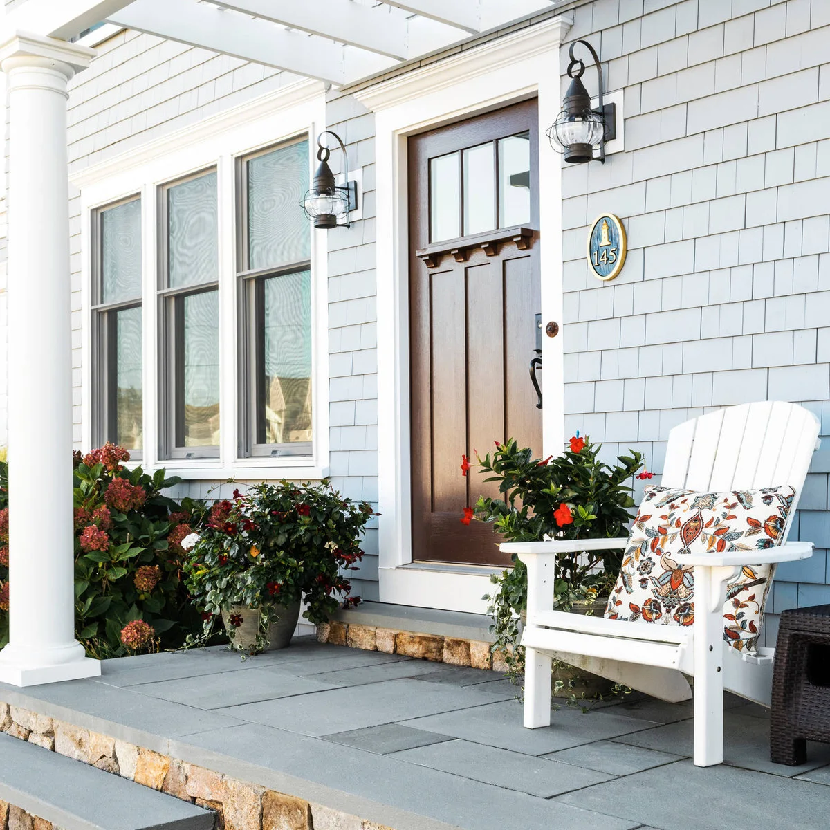 A white porch chair sits beside a brown front door, pale shingle siding, paired black lanterns and flowering containers.