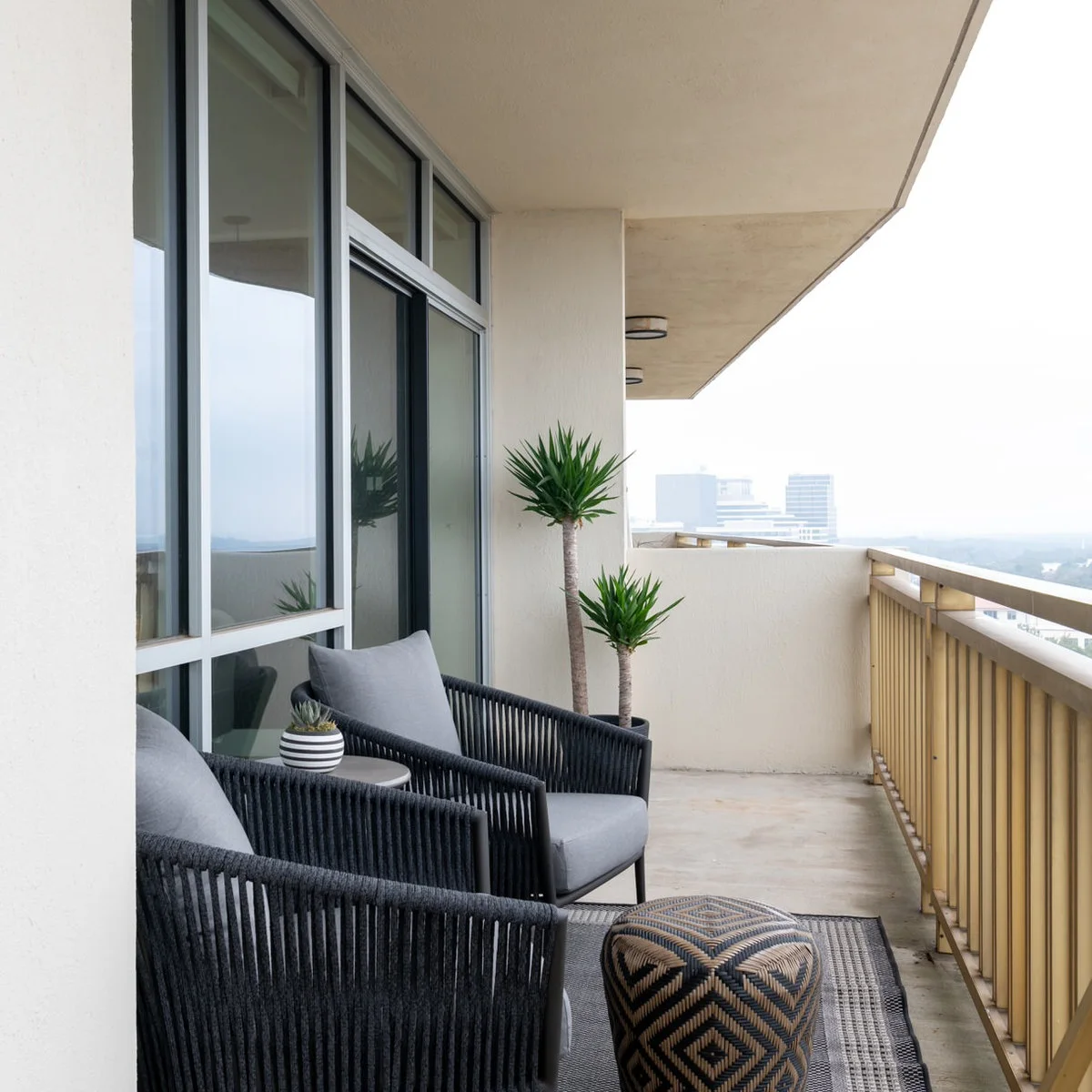 Two dark corded chairs and a patterned pouf sit along the wall of a narrow pale balcony