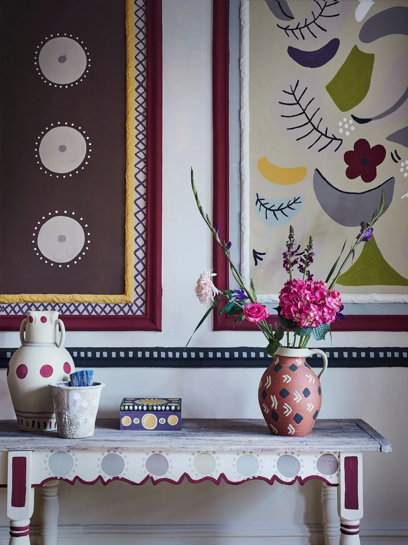 Decoratively painted country console with dotted edges, colorful patterned wall panels and bright flowers in a decorated jug