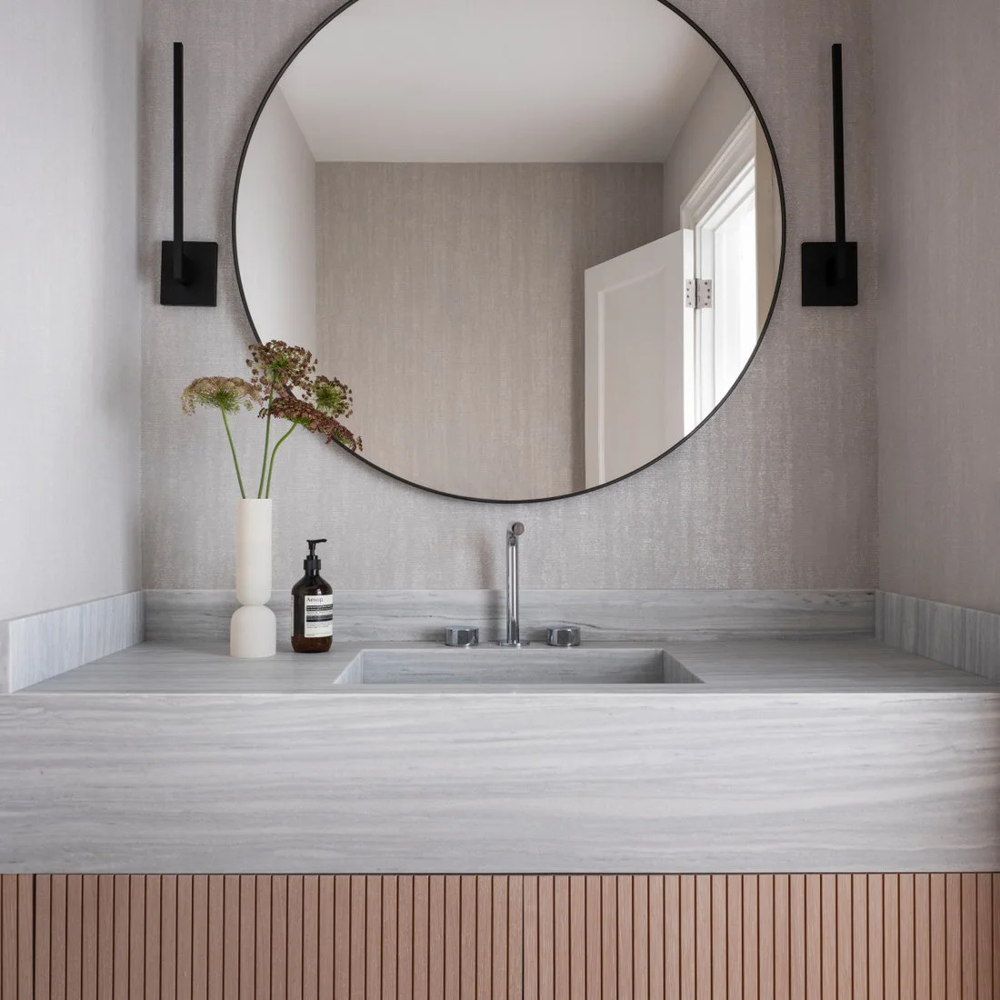 A wood slatted vanity front sits below a pale gray veined counter, broad round mirror and two slim black vertical lights against textured neutral walls.