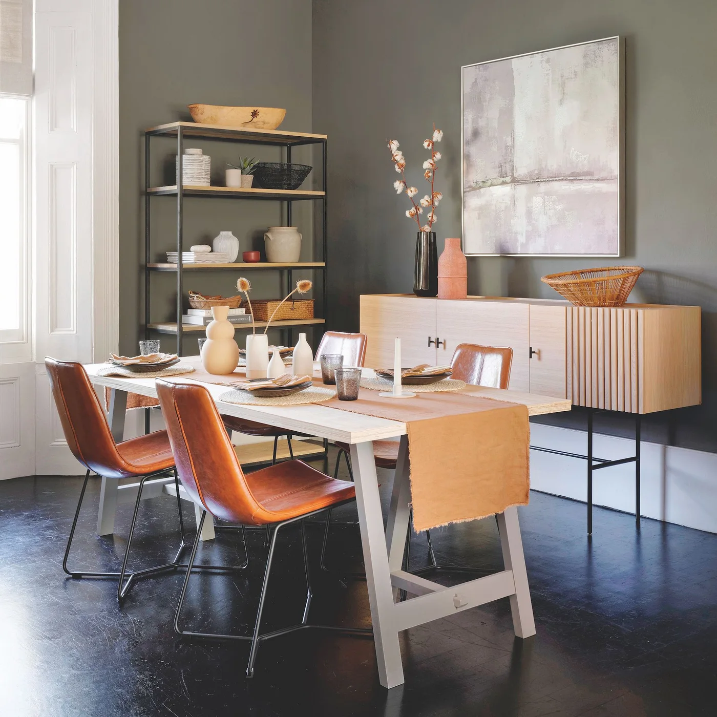 Muted green walls frame a pale wooden table, warm brown chairs, open shelving and a light wood sideboard