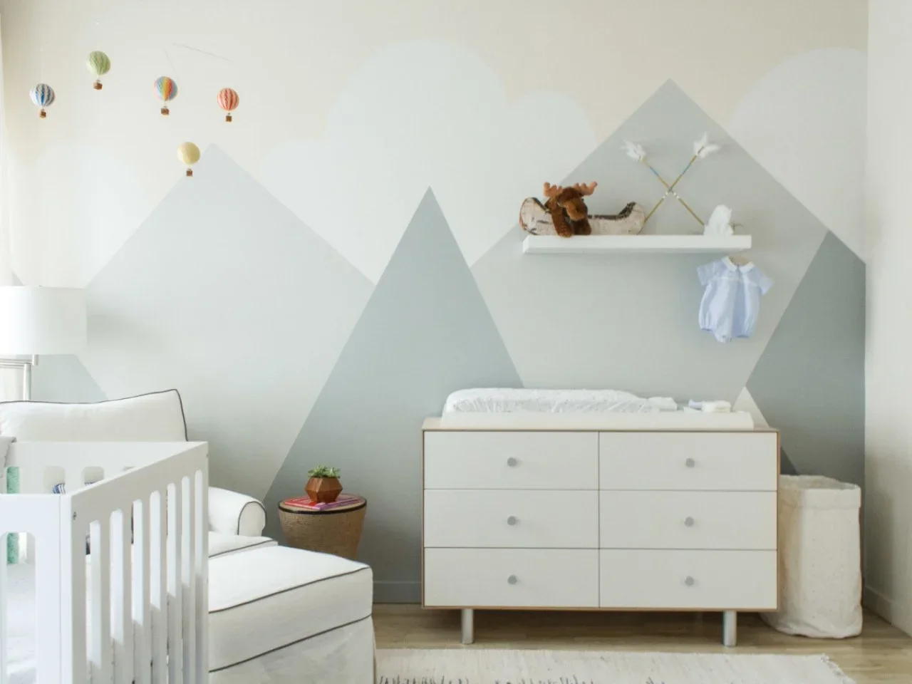 A pale mountain mural rises behind a white changing chest beside an adult chair and a small round side table.