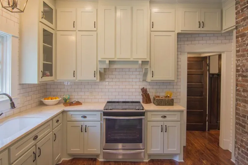 White framed kitchen cabinets with pale rectangular backsplash tiles extending around a small doorway and exposed brick at the side.
