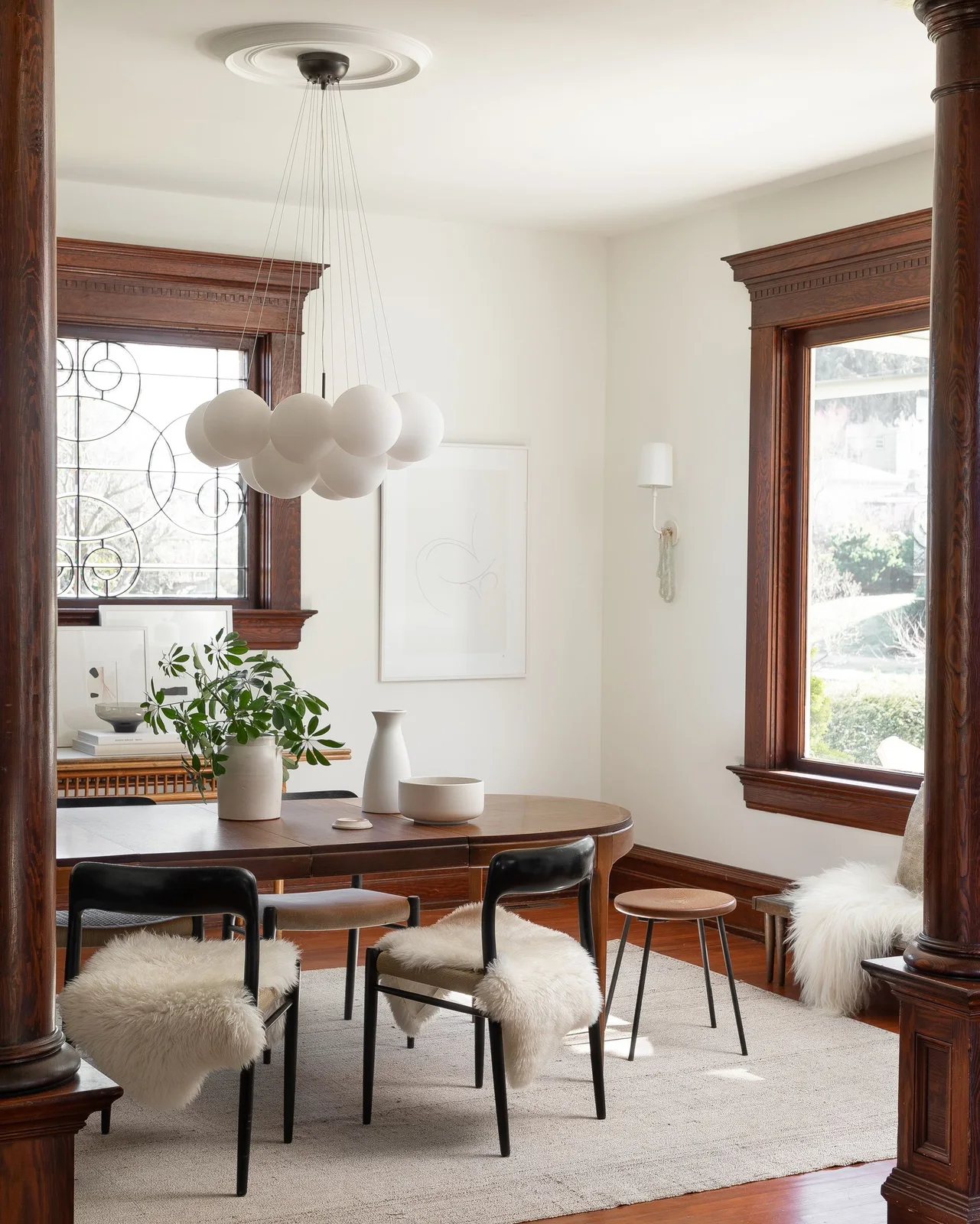 A wood dining table and dark chairs with pale shaggy seat coverings stand beneath clustered white globe lights beside white walls and dark wood window trim