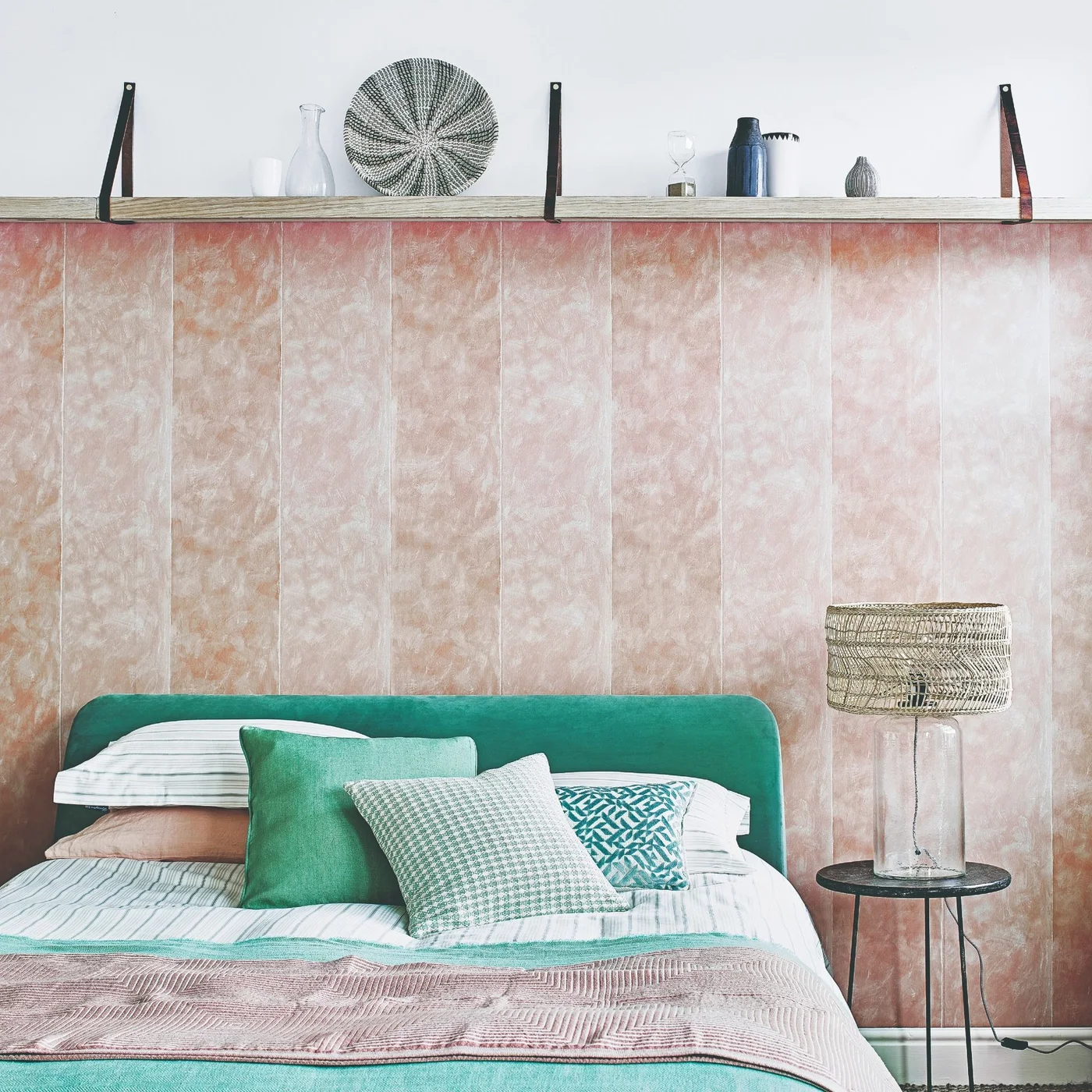 Pink marbled panel-effect wallpaper rises behind a teal bed below a timber display shelf.