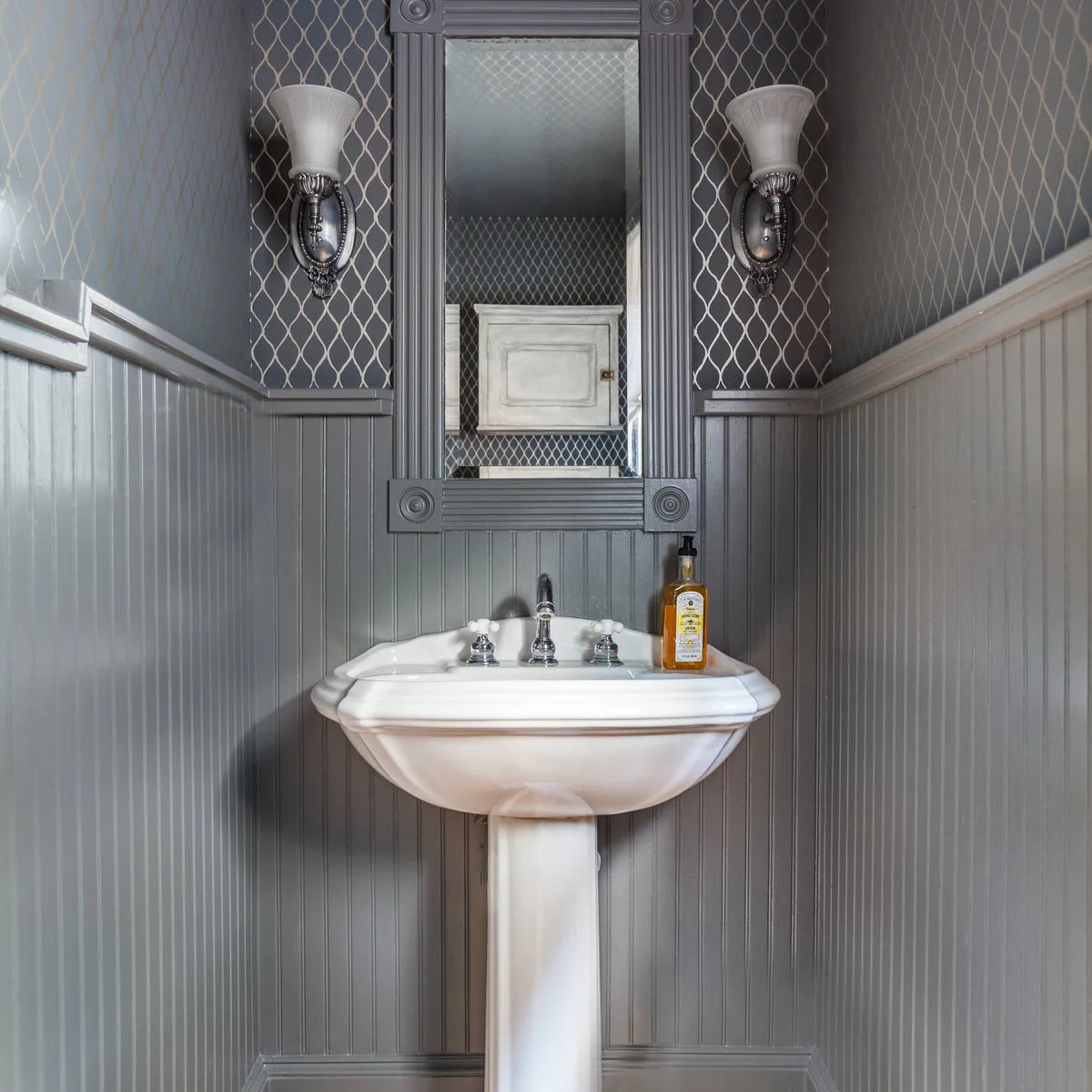 A white pedestal sink sits in a narrow gray-paneled alcove below a tall framed mirror and patterned upper walls.