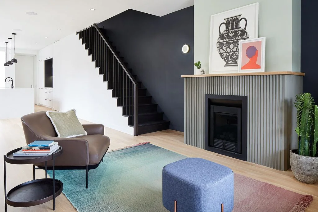 A straight black fireplace opening sits within a gray ribbed surround under a pale wood mantel beside dark stairs, a pale green wall and a muted multicolored rug