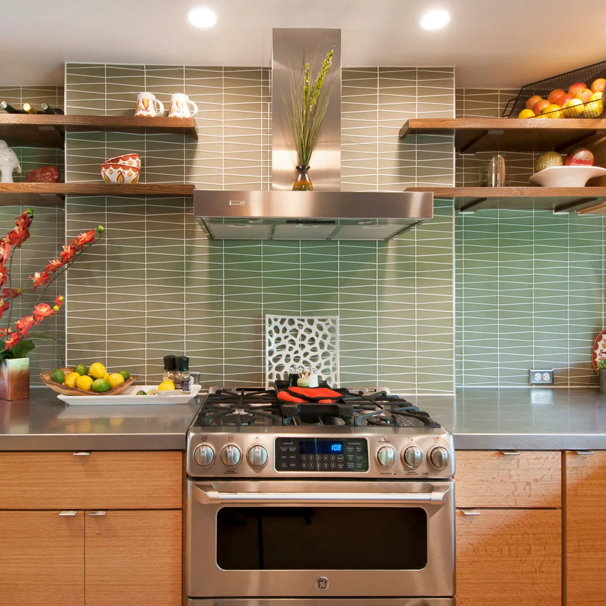 Wood lower cabinets and open shelves around a metal hood, against green geometric tiles with pale grout lines.