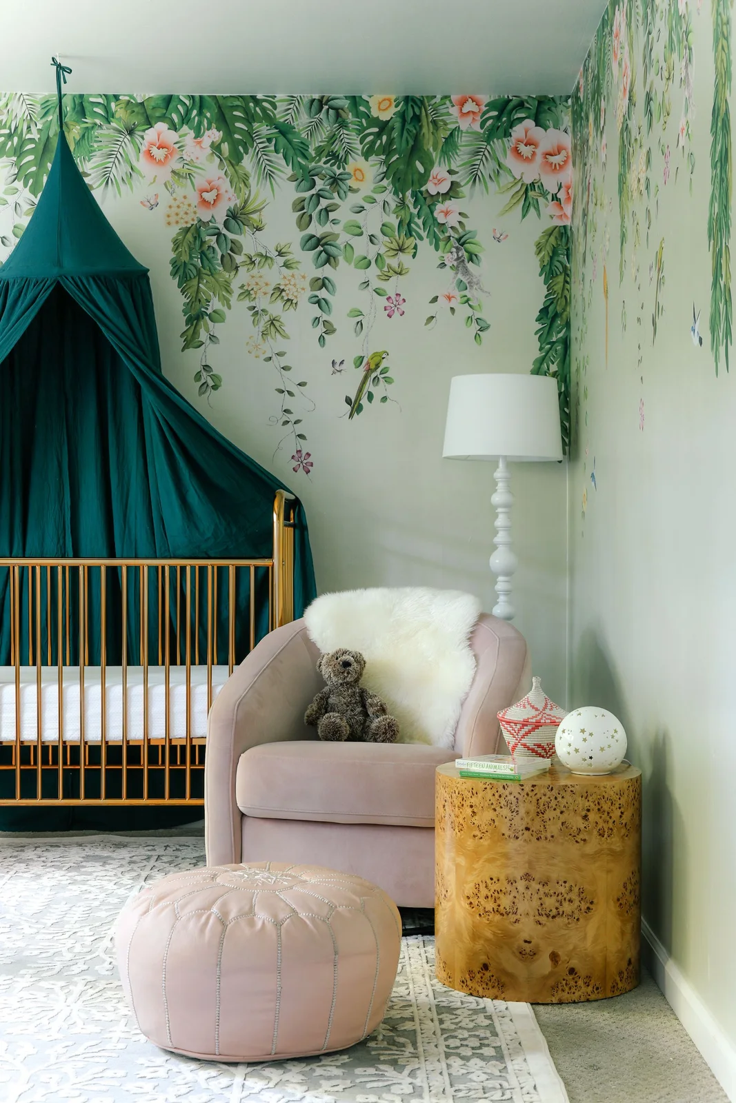 A leafy floral nursery mural surrounds a gold-colored crib with a green canopy beside a pale pink adult chair, round pouf and yellow side table