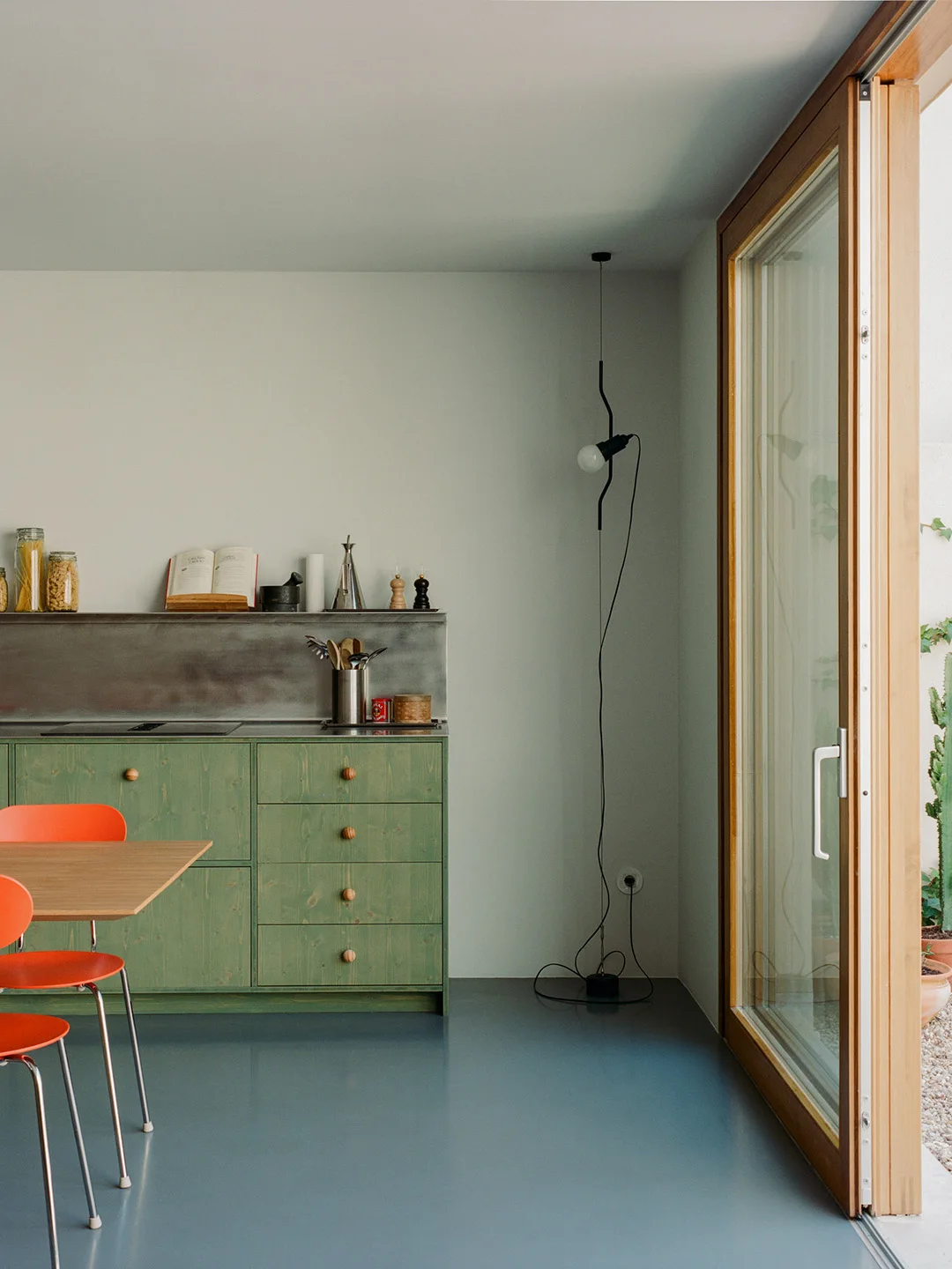 Muted green lower cabinets with small round wooden knobs sit beneath a gray backsplash beside an orange chair and glazed door