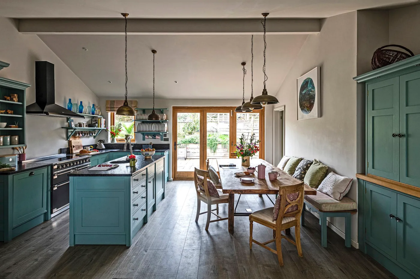 Kitchen with teal cabinets and island, wooden dining table, cushioned bench and glazed garden doors.
