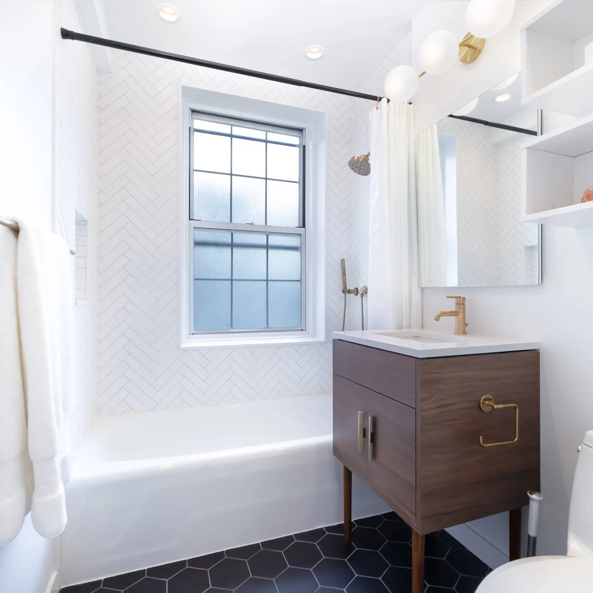 A plain white shower curtain hangs from a black rod beside herringbone wall tile, a wood-toned vanity and dark hexagonal floor tiles.