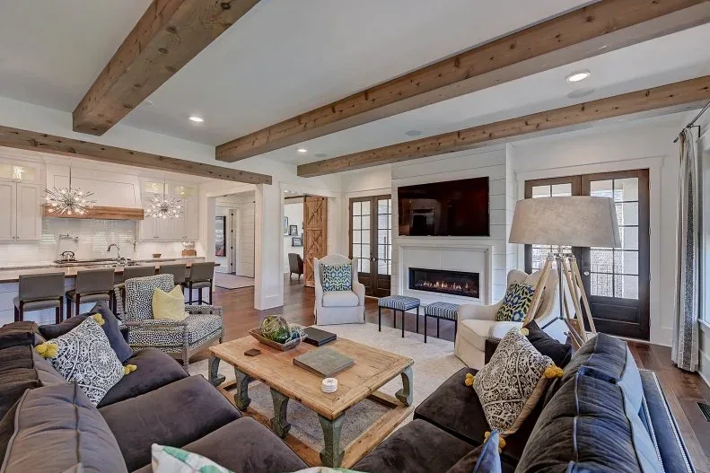 Open farmhouse room with dark sofas, wood table, pale chairs, exposed beams and fireplace wall beside a white kitchen island