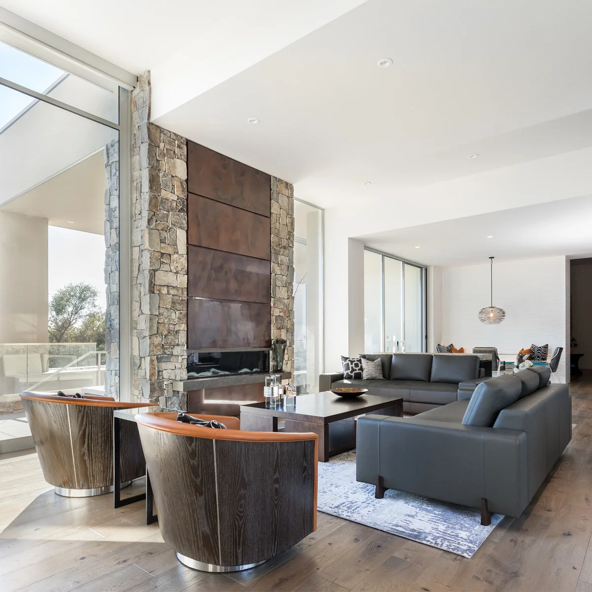 Charcoal sofas face curved chairs with cognac-colored upholstery beside a tall mixed-tone fireplace wall.