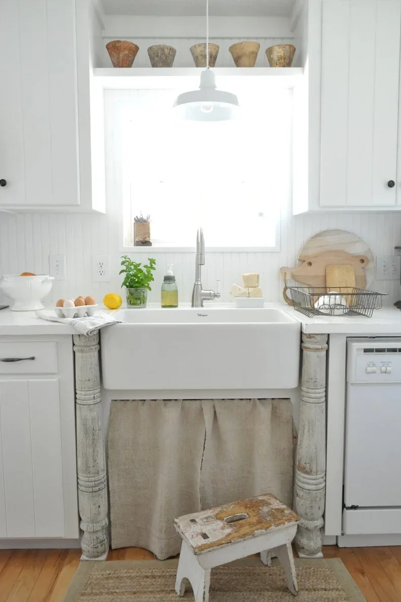 White apron-front sink between pale detailed supports, with gathered neutral curtain below and a bright window above