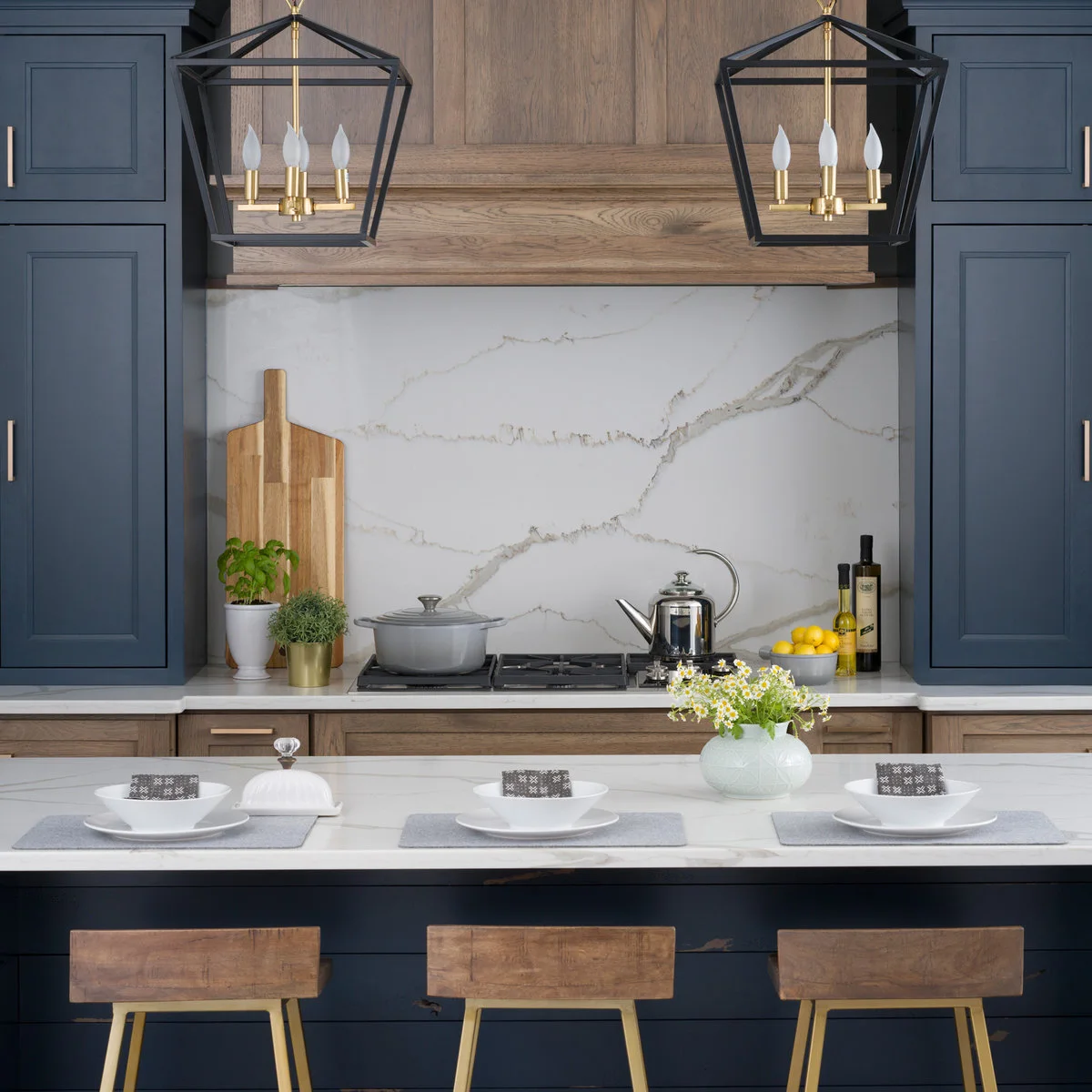 Two dark open lantern frames with warm metal candle-style holders hang above a navy island beside blue cabinets and a wood hood.