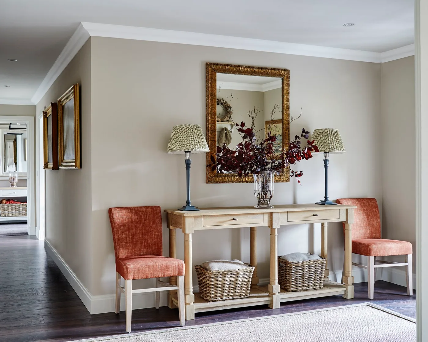 Pale wood console with central red foliage, two blue lamps, gold-framed mirror, orange chairs and baskets on the lower shelf