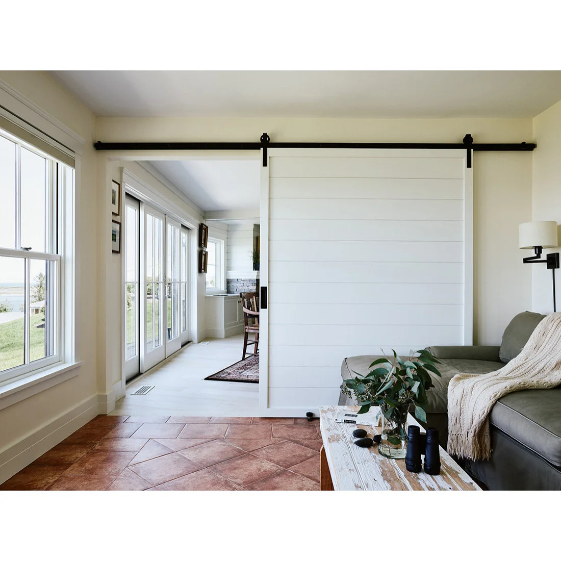 A terracotta floor has diagonal center tiles and a straight border beside a gray sofa and white sliding door.